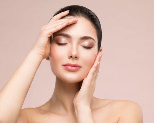 Beautiful Woman Face Skin Care. Natural Make up. Skin Health Treatment. Model massaging Face by Hands. Beige Studio background. Fashion Girl with Closed eyes