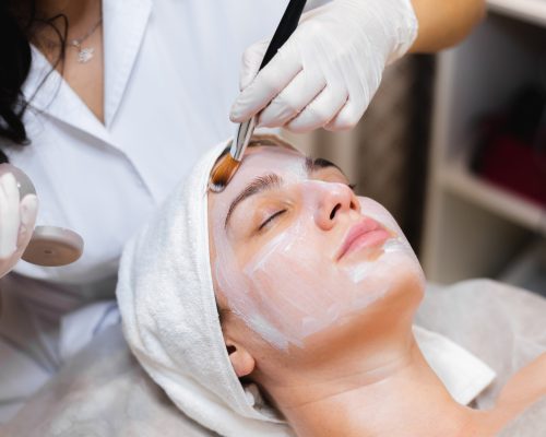 Beautician with a brush applies a white moisturizing mask to the face of a young girl client in a spa beauty salon